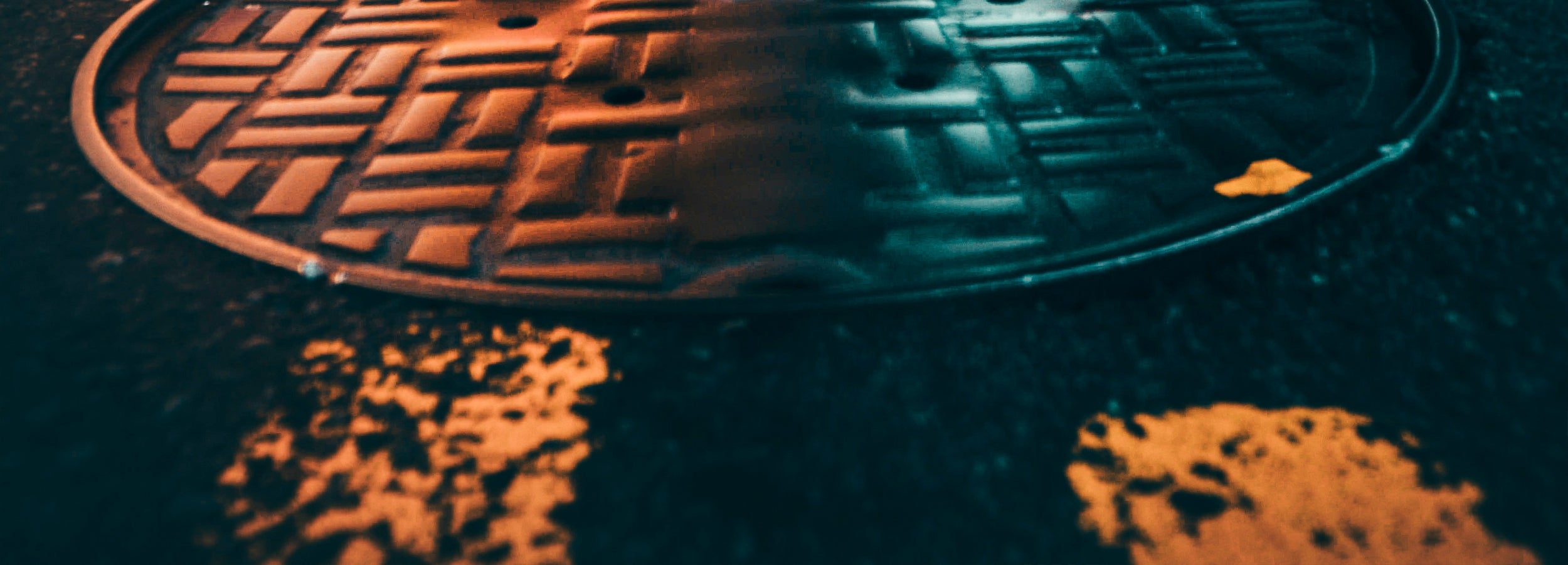 A close-up, angled view of a round metal manhole cover on dark asphalt, with orange streetlights reflecting on the wet surface.