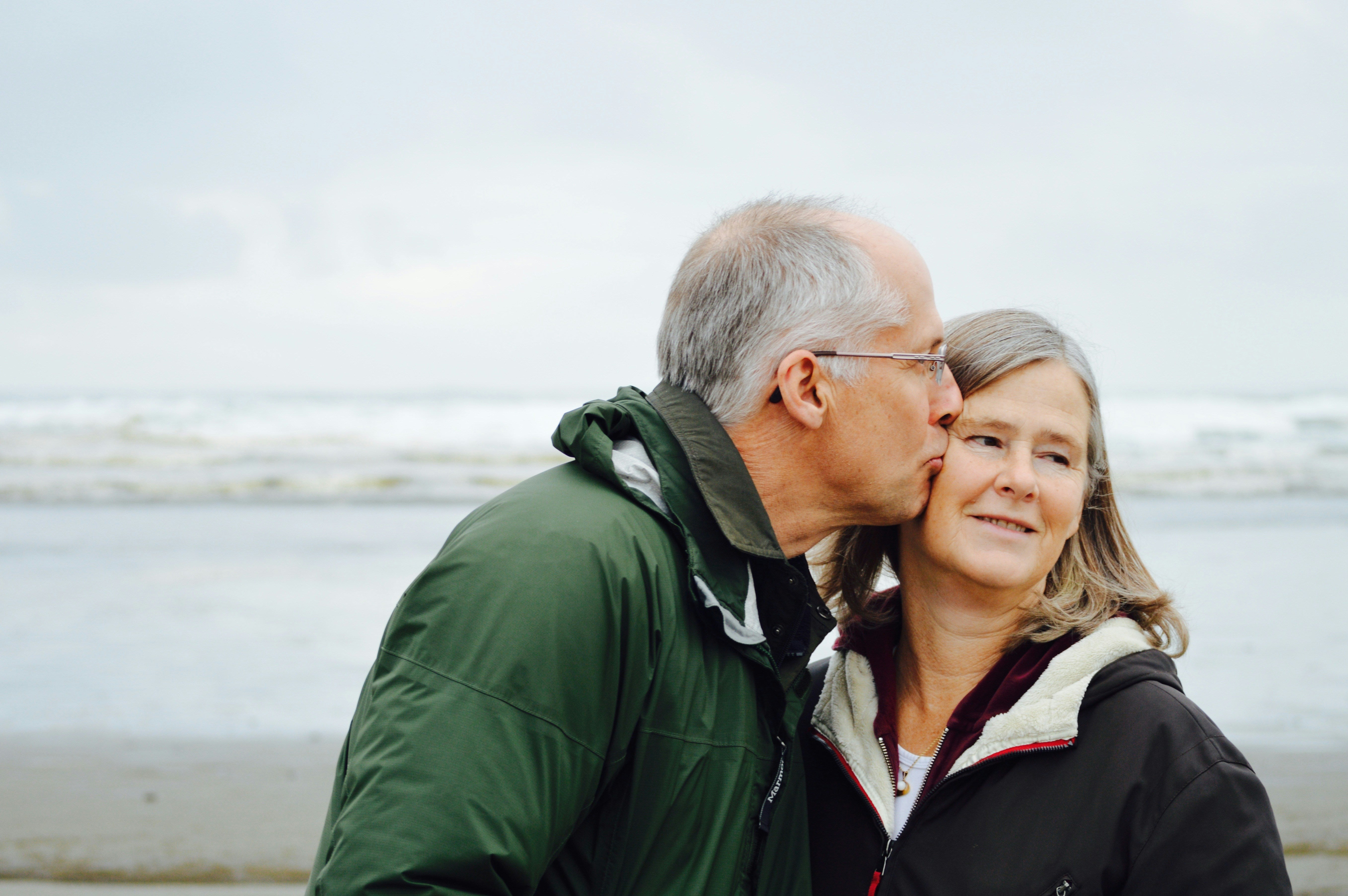 Man and woman standing close together on a beach with water in the background. The man is kissing the woman's cheek.