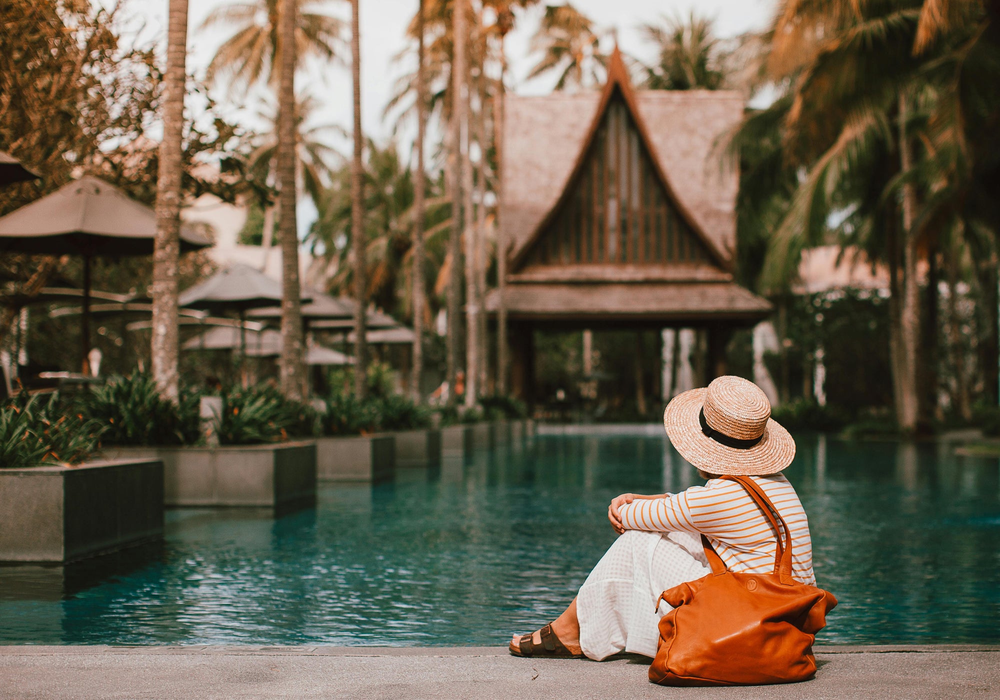 Woman sitting by a pool with palm trees and a wooden hut in the background.