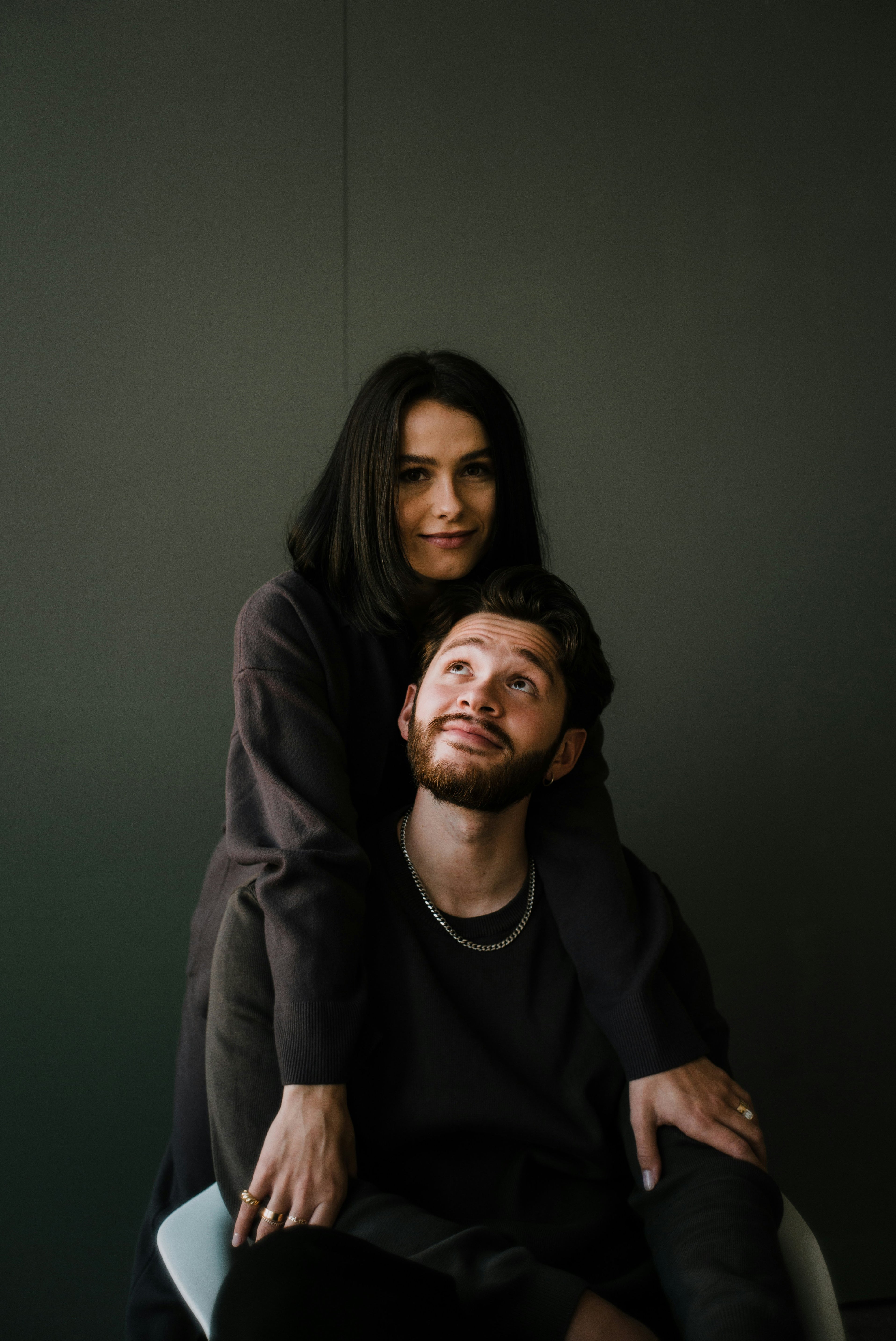 A man and a woman in dark clothing pose together against a dark gray background. The woman is seated behind the man, who is looking upward, with both wearing black or very dark charcoal tops and bottoms.