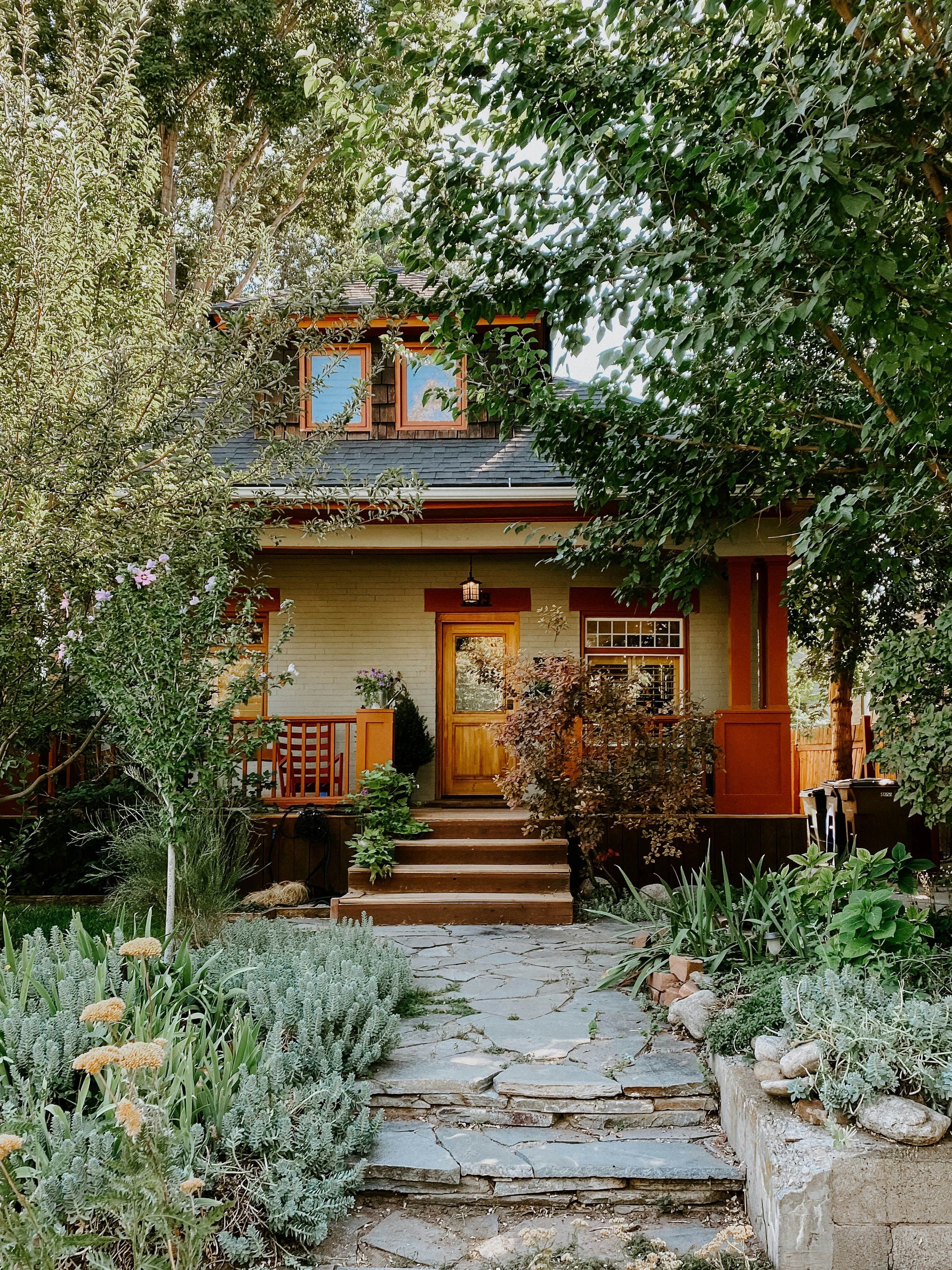 A charming cottage-style house with a stone path leading up the front steps, surrounded by lush green garden plants and trees. The house features a light beige exterior, warm orange-brown trim, a wooden front door, and a dark gray roof.