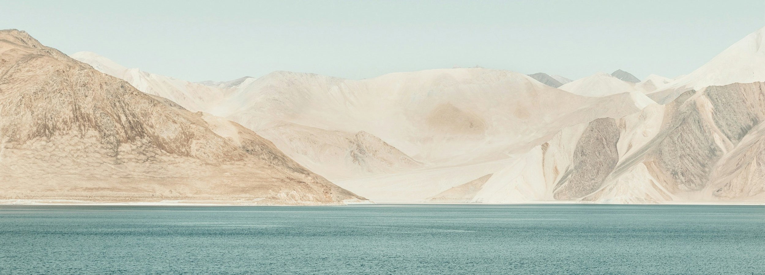 A scenic landscape print featuring a calm blue lake in the foreground and large, pale beige and brown mountains under a soft light blue sky.