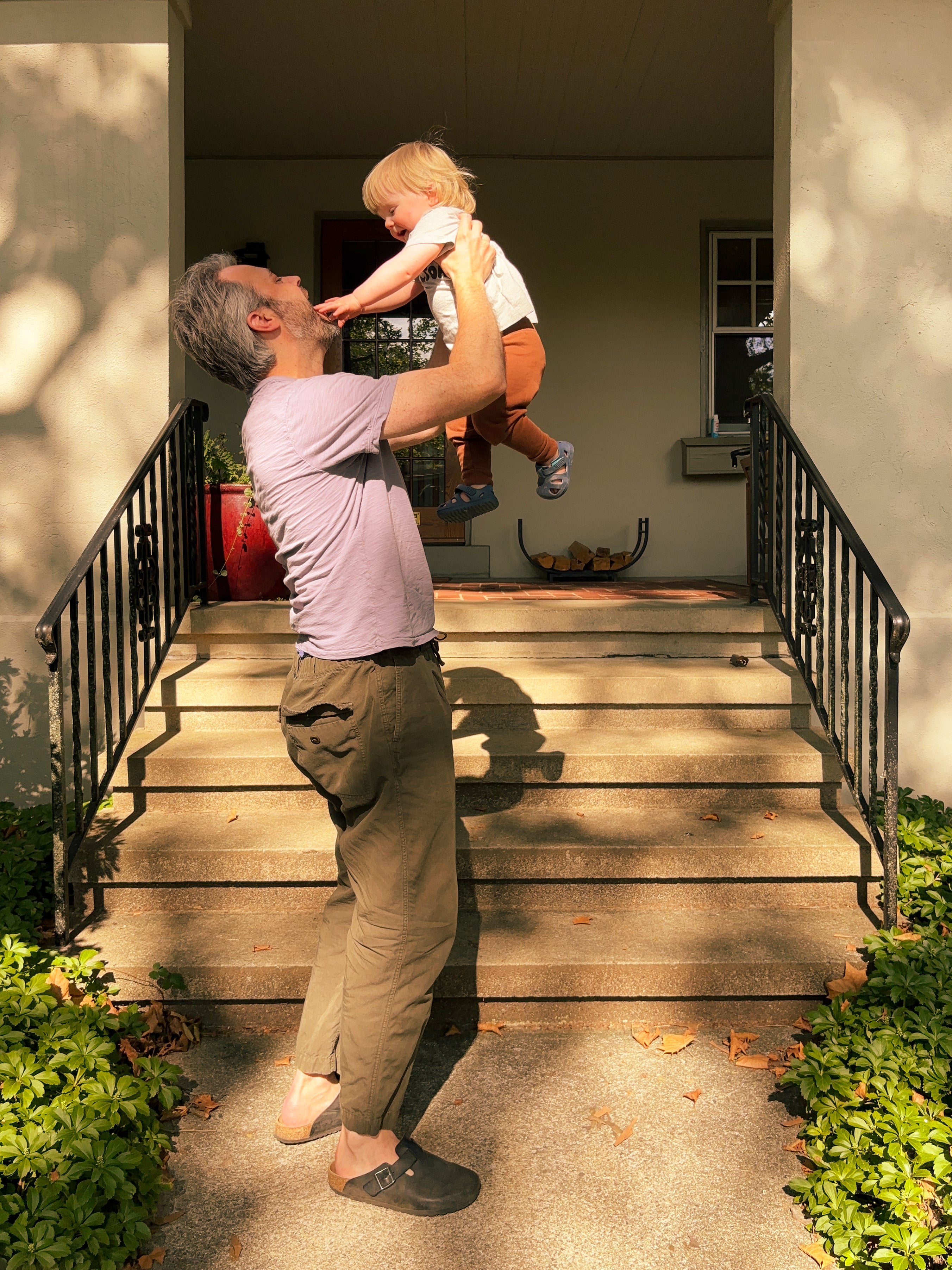A man in a lavender t-shirt and khaki pants holds a young child up in the air on a sunny porch with concrete steps and black metal railings.