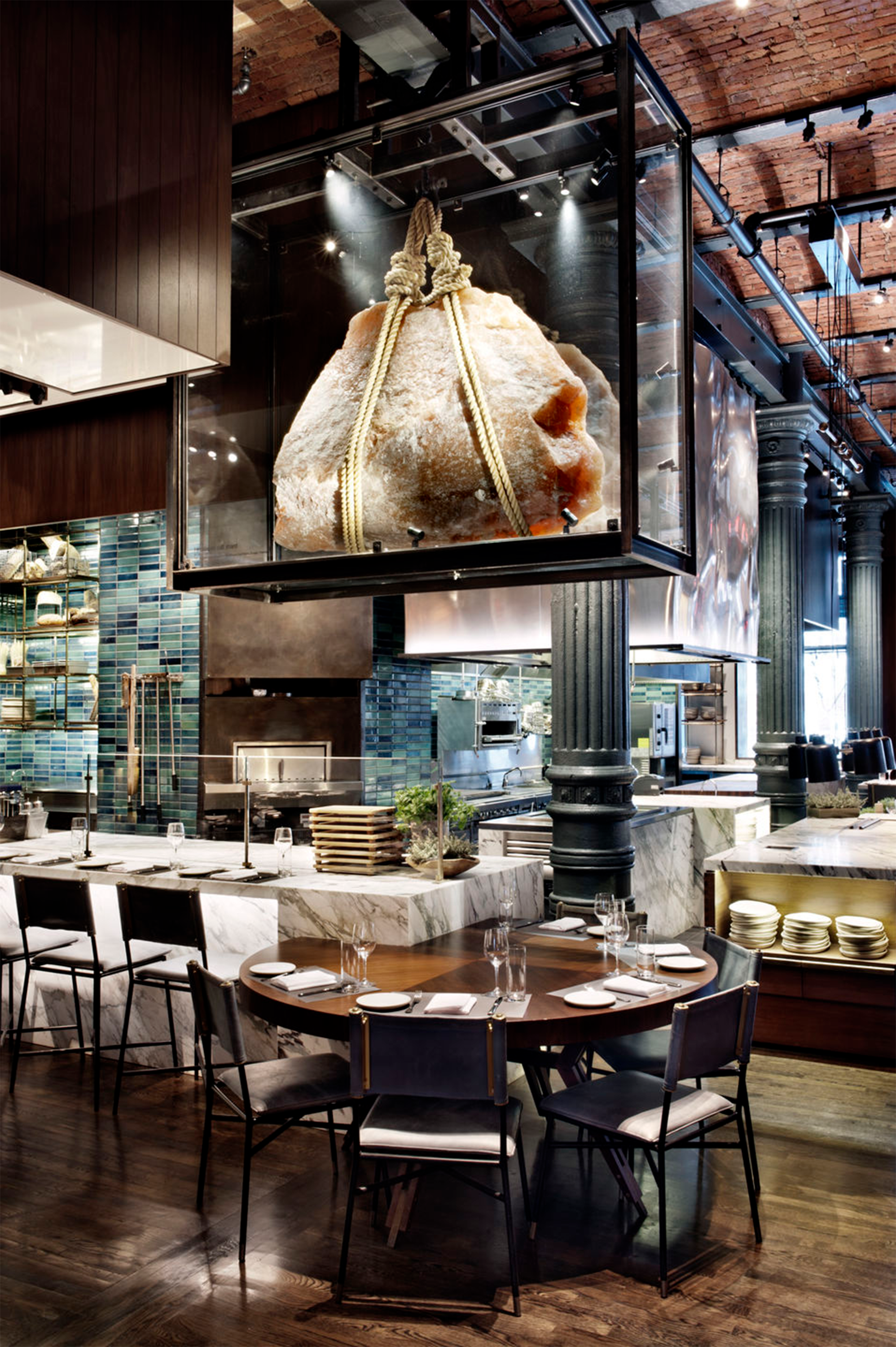 A large Himalayan salt rock is suspended from the ceiling in a restaurant setting. The salt rock is encased in a glass box. The open kitchen is seen in the background.