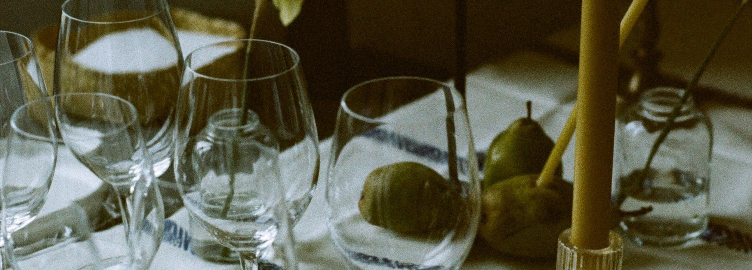 A close-up, slightly soft shot of a table setting with several clear wine glasses, a lit yellow candle in a glass holder, and green pears on a white tablecloth.