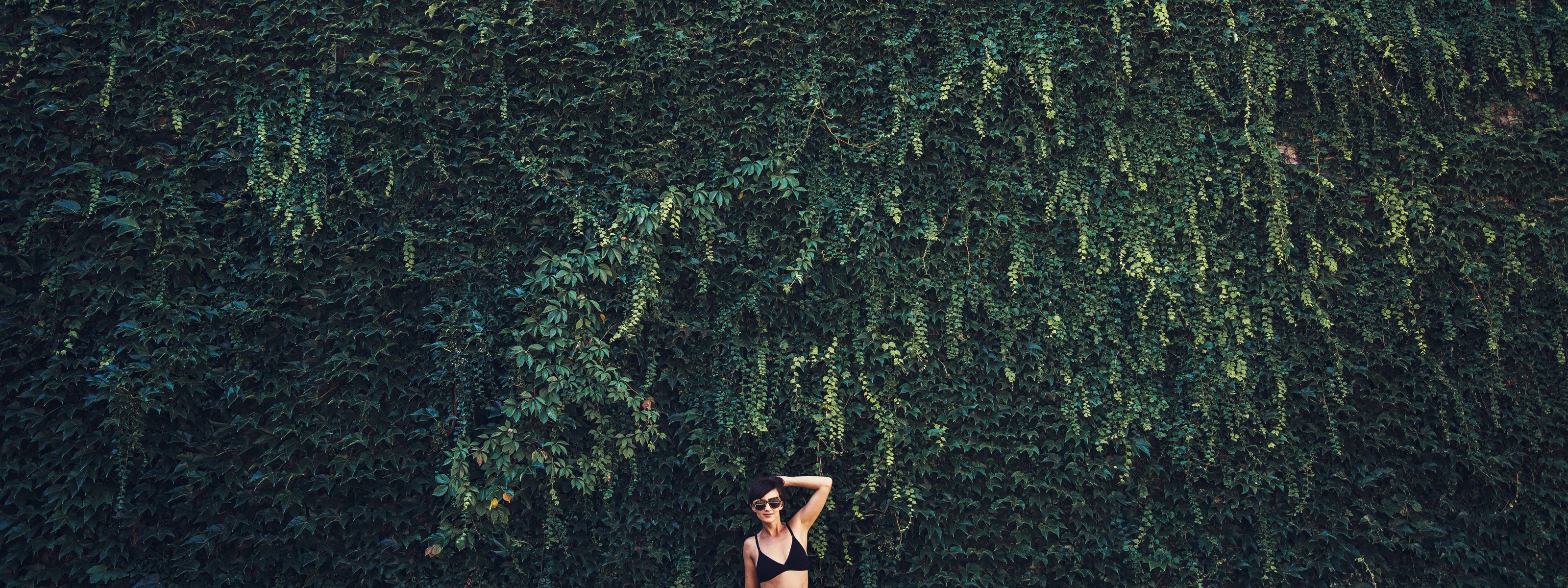 A woman poses in front of a large wall covered with ivy.