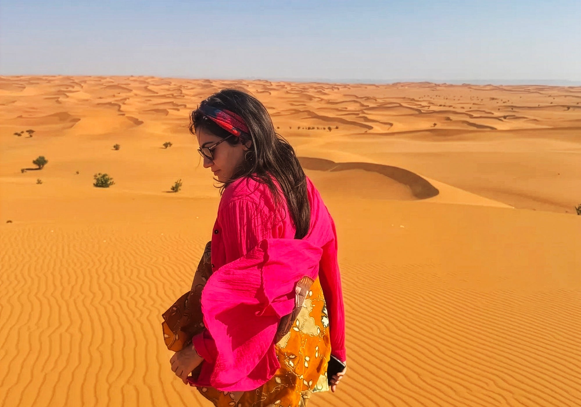 A woman wearing a bright pink traditional outfit and sunglasses walks across golden sand dunes in a desert.landscape.